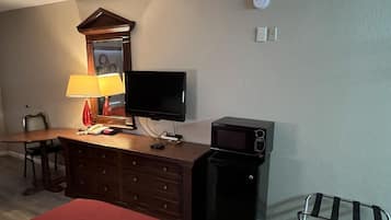 A hotel room with a wooden desk, a television, a microwave, a red bedspread, a dresser with drawers, a lamp, a chair, and a small table with a red tablecloth.