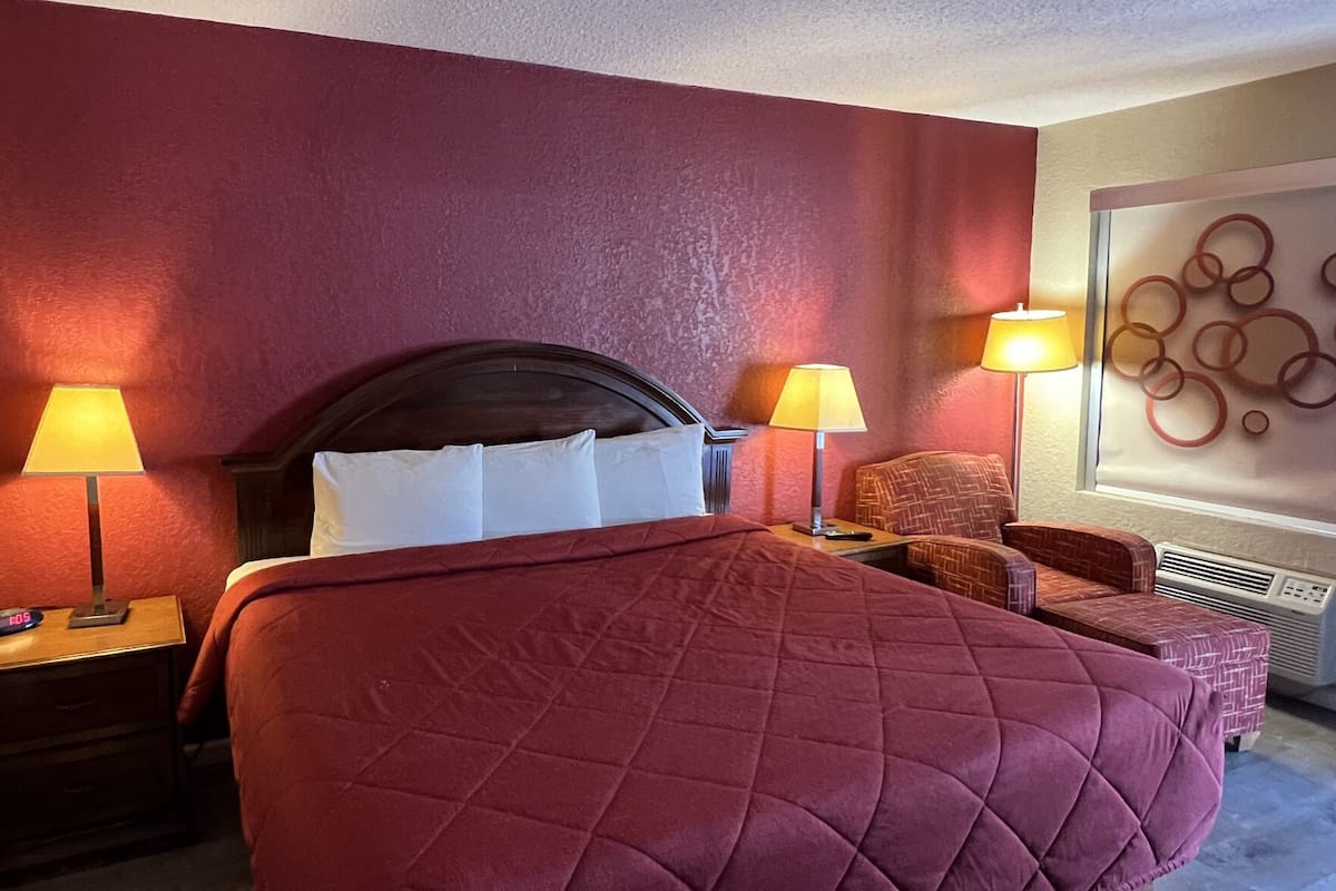 A hotel room with a large bed, two bedside tables with lamps, a chair, a wall art piece, and a window with blinds.