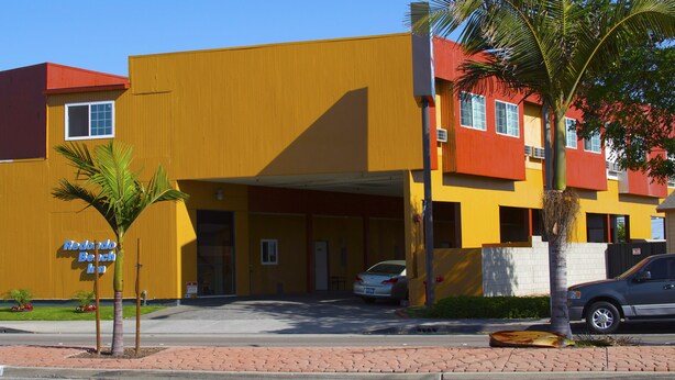 A yellow building with a red roof, a palm tree, and a car parked in front. - Redondo Beach Inn (Gardena)