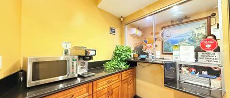 A kitchen with a microwave, coffee machine, and wooden cabinets. There is a sign on the window indicating maximum occupancy and a face mask requirement.