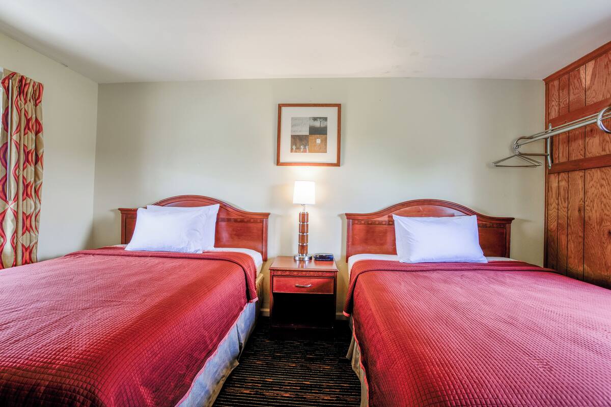 A hotel room with two beds, a nightstand with a lamp, a wooden wardrobe, and a framed picture on the wall.