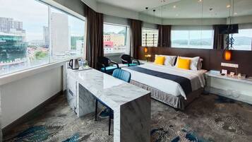 A hotel room with a bed, desk, chairs, and a large window offering a city view.