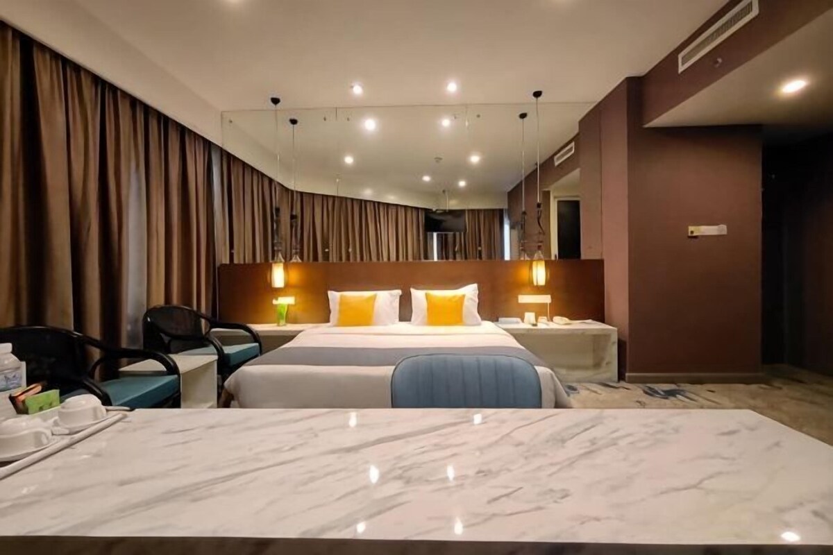 A hotel room with a bed, a desk, a chair, a nightstand, and a bathroom mirror.