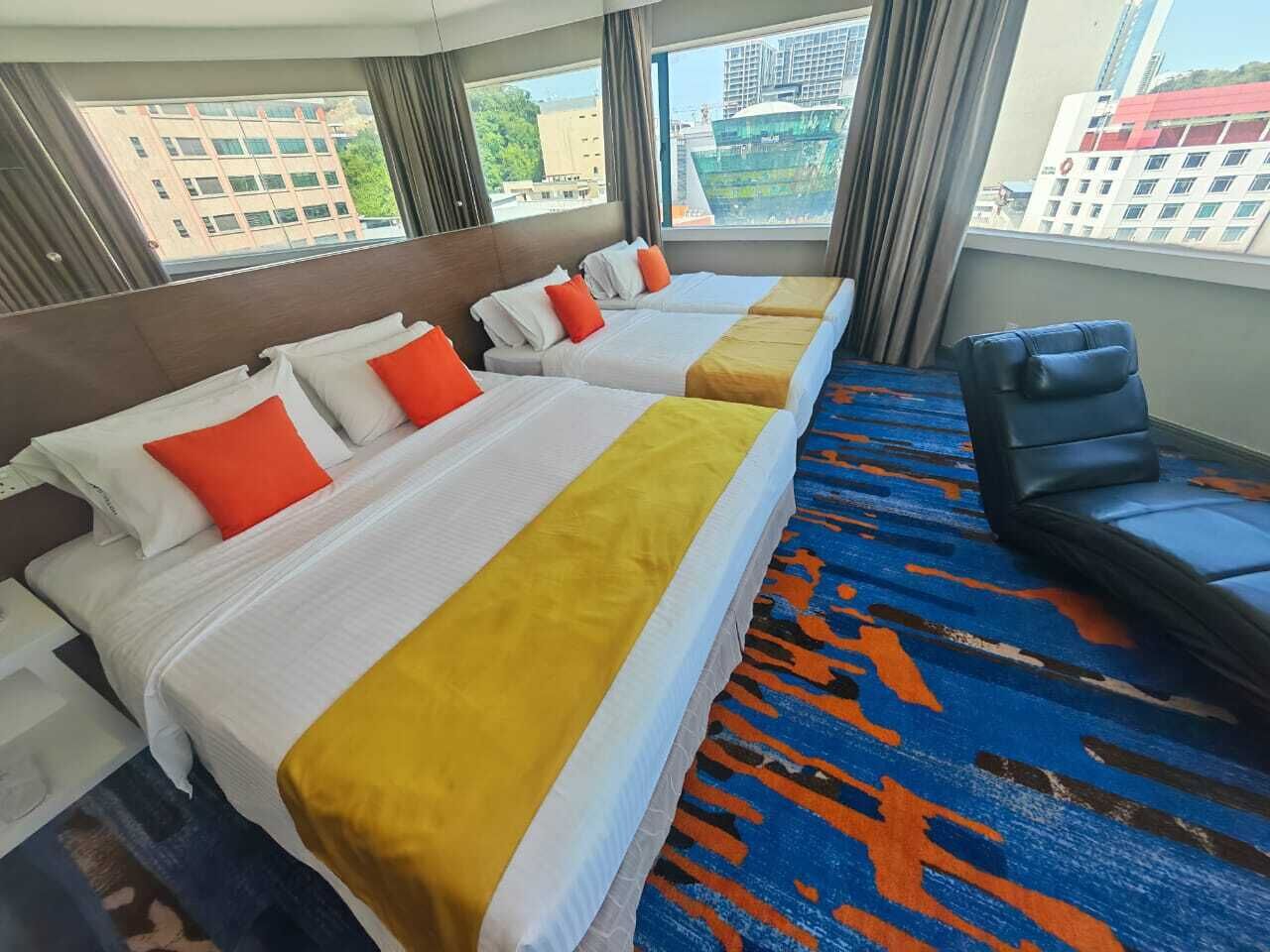 Une chambre d’hôtel avec deux lits, une chaise bleue et une vue sur les immeubles à travers de grandes fenêtres.