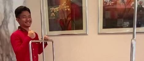 Uma pessoa de uniforme vermelho empurra um carrinho com um logotipo em um quarto com parede de mármore e quadros.