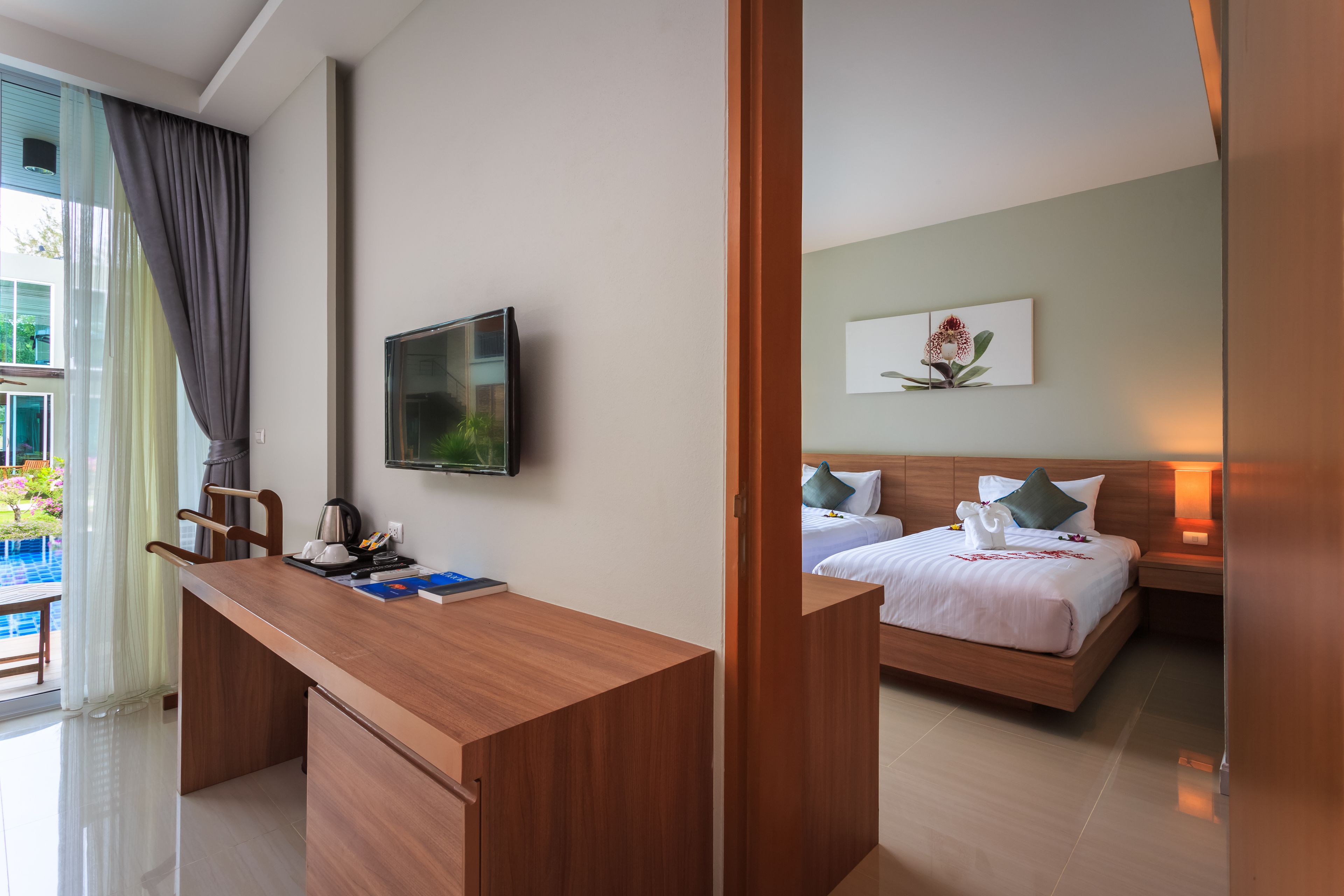 A hotel room with a wooden desk, a bed with a white comforter, a flat-screen TV mounted on the wall, and a painting above the bed.
