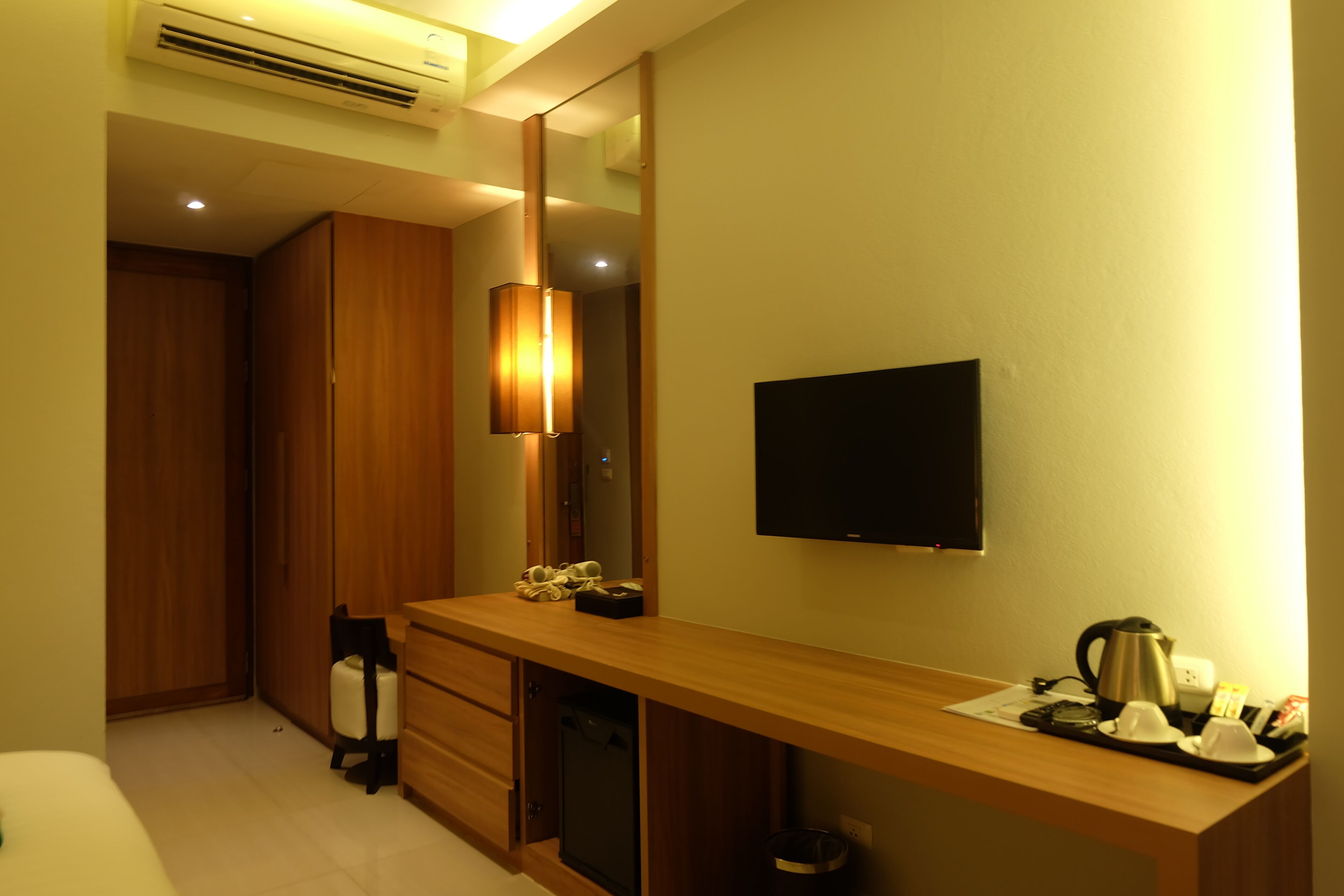 A hotel room with a wooden desk, a television, a mirror, a wardrobe, and a small kitchenette area.