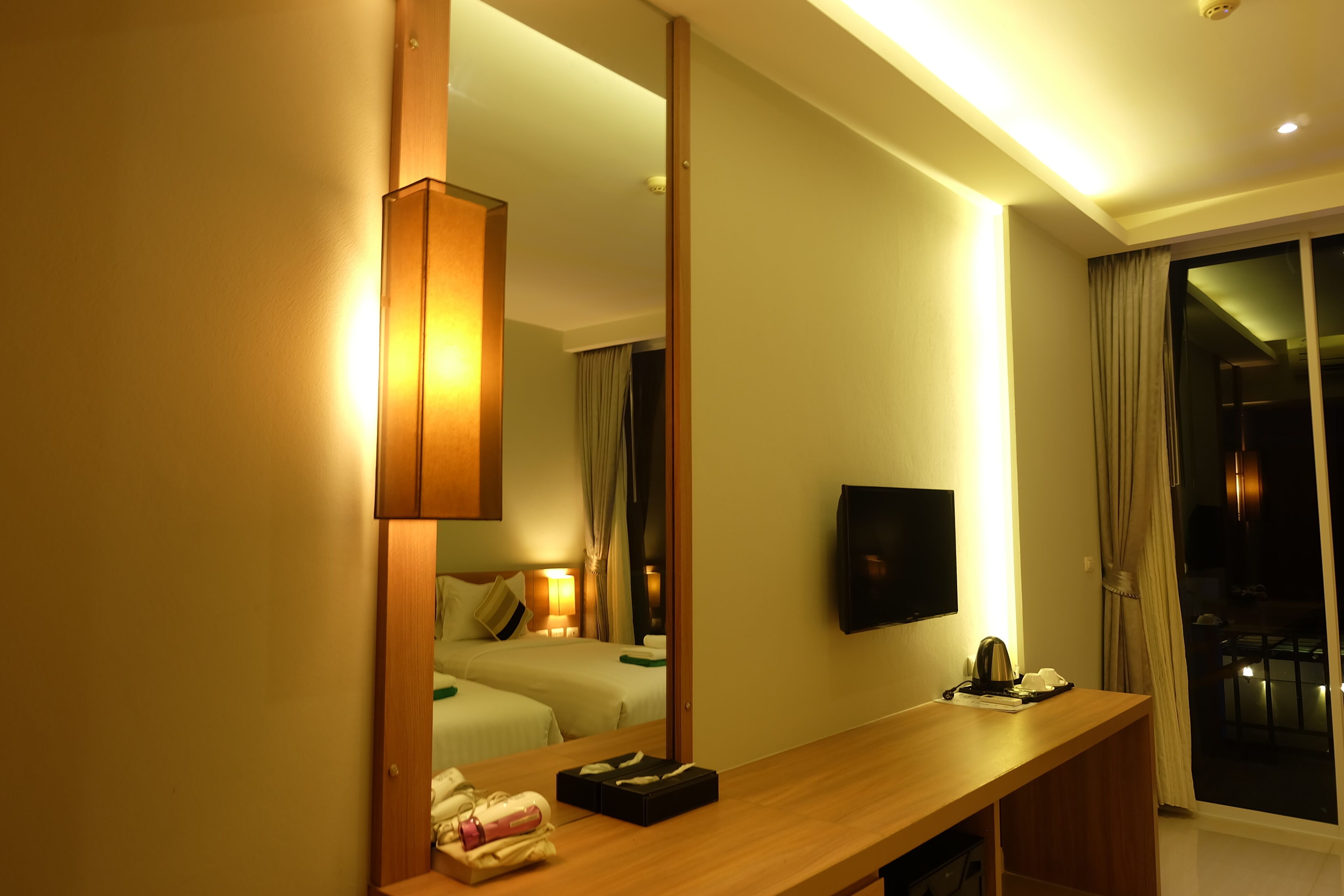 Habitación de hotel con un espejo grande, un escritorio de madera, un televisor de pantalla plana y un balcón con cortinas.
