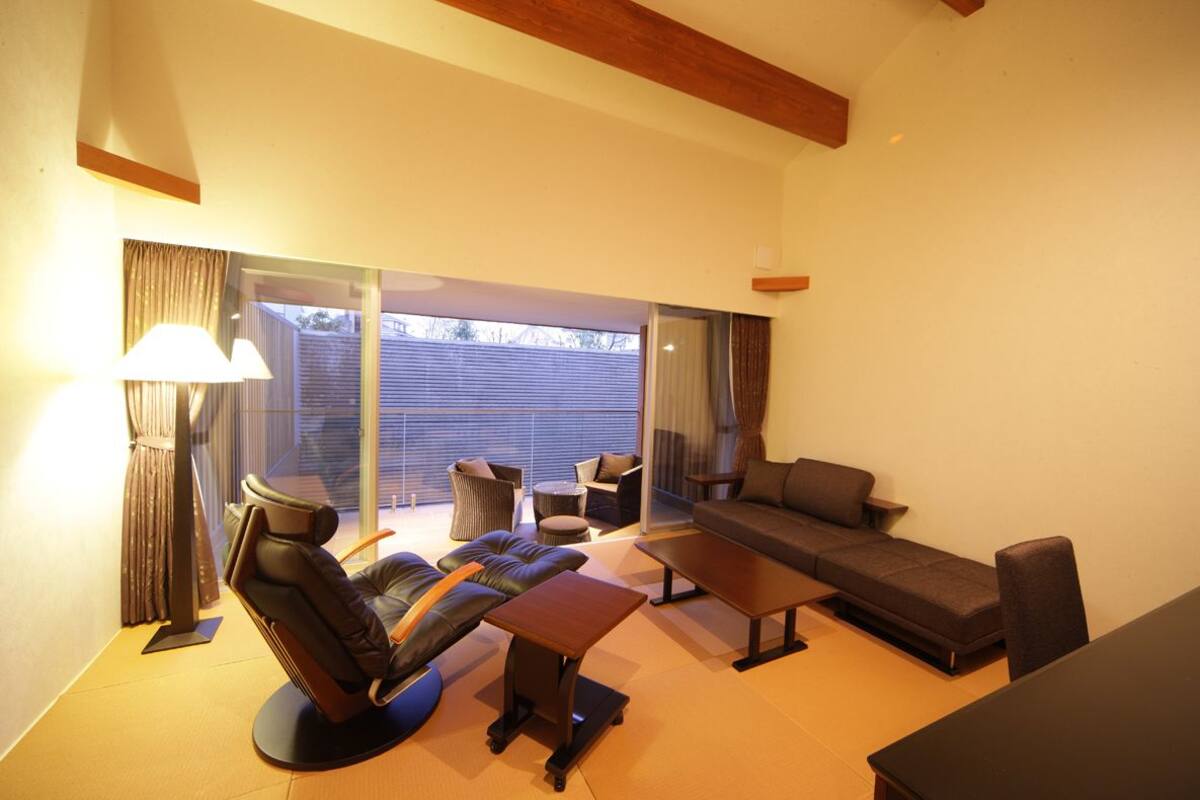 Een moderne woonkamer met een bank, stoelen, een salontafel en een massagestoel. Er is een schuifpui die leidt naar een buitenruimte.