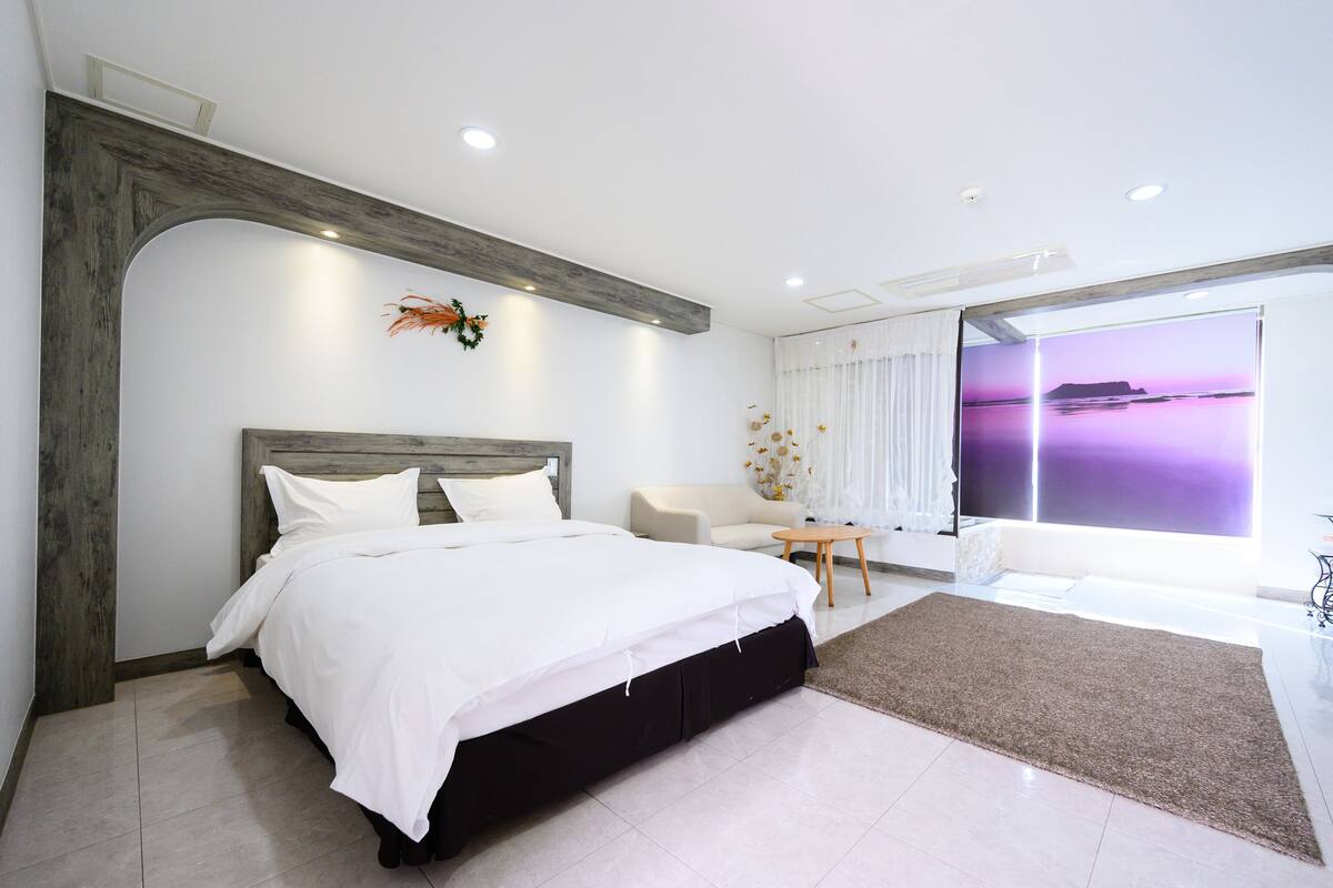 A modern bedroom with a large bed, a wooden headboard, a small table, and a view of the sea through a large window.