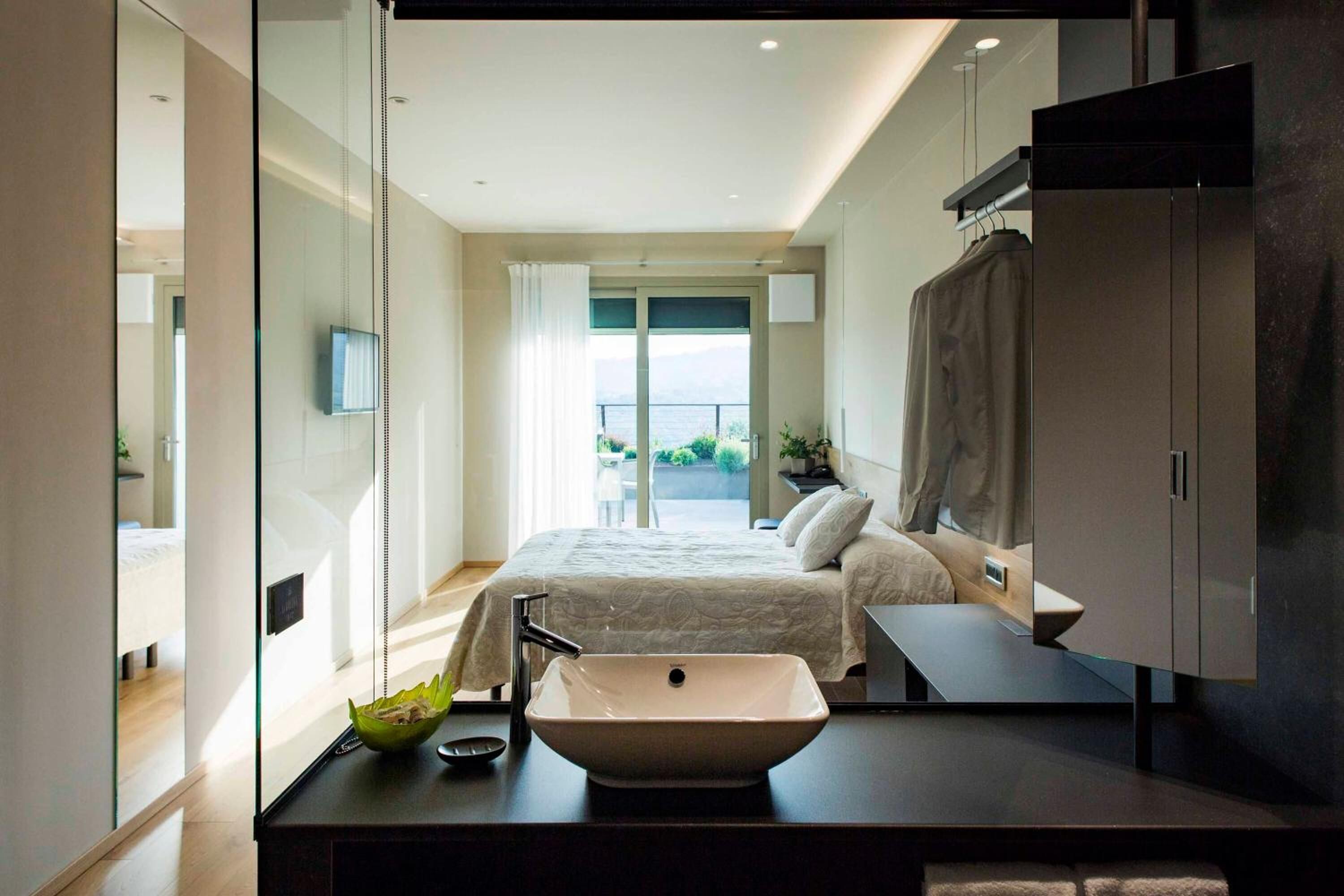 A modern bedroom with a large bed, a bedside table with a sink, and a view of the outdoors.