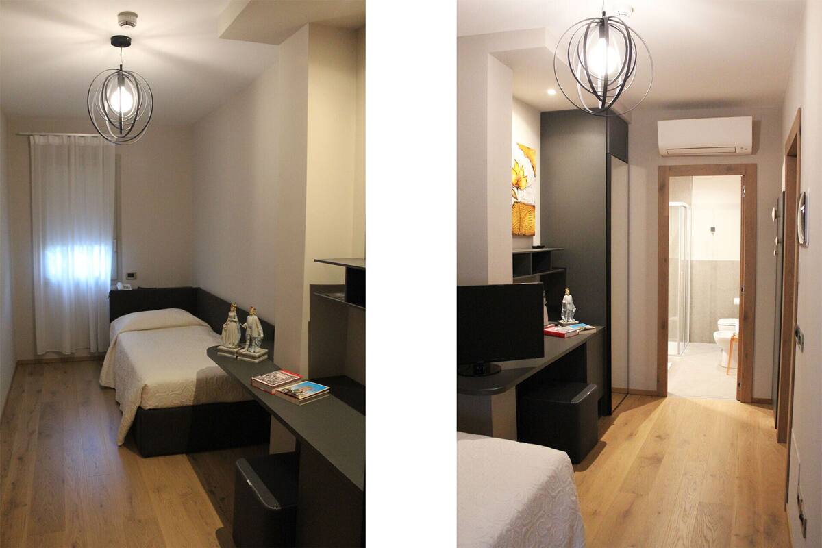 A modern hotel room with a bed, bedside table, and a desk with a chair. There is a ceiling light fixture, a window with curtains, and an air conditioning unit.