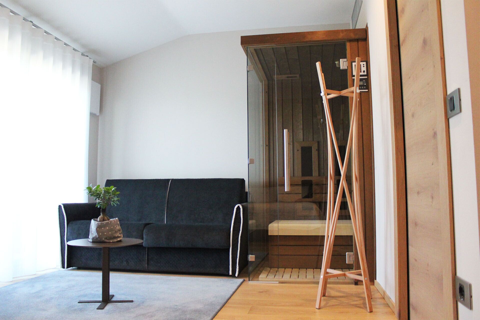 Una sala moderna con un sofá azul oscuro, una mesita redonda con una planta y una escalera de madera apoyada en una puerta de cristal.