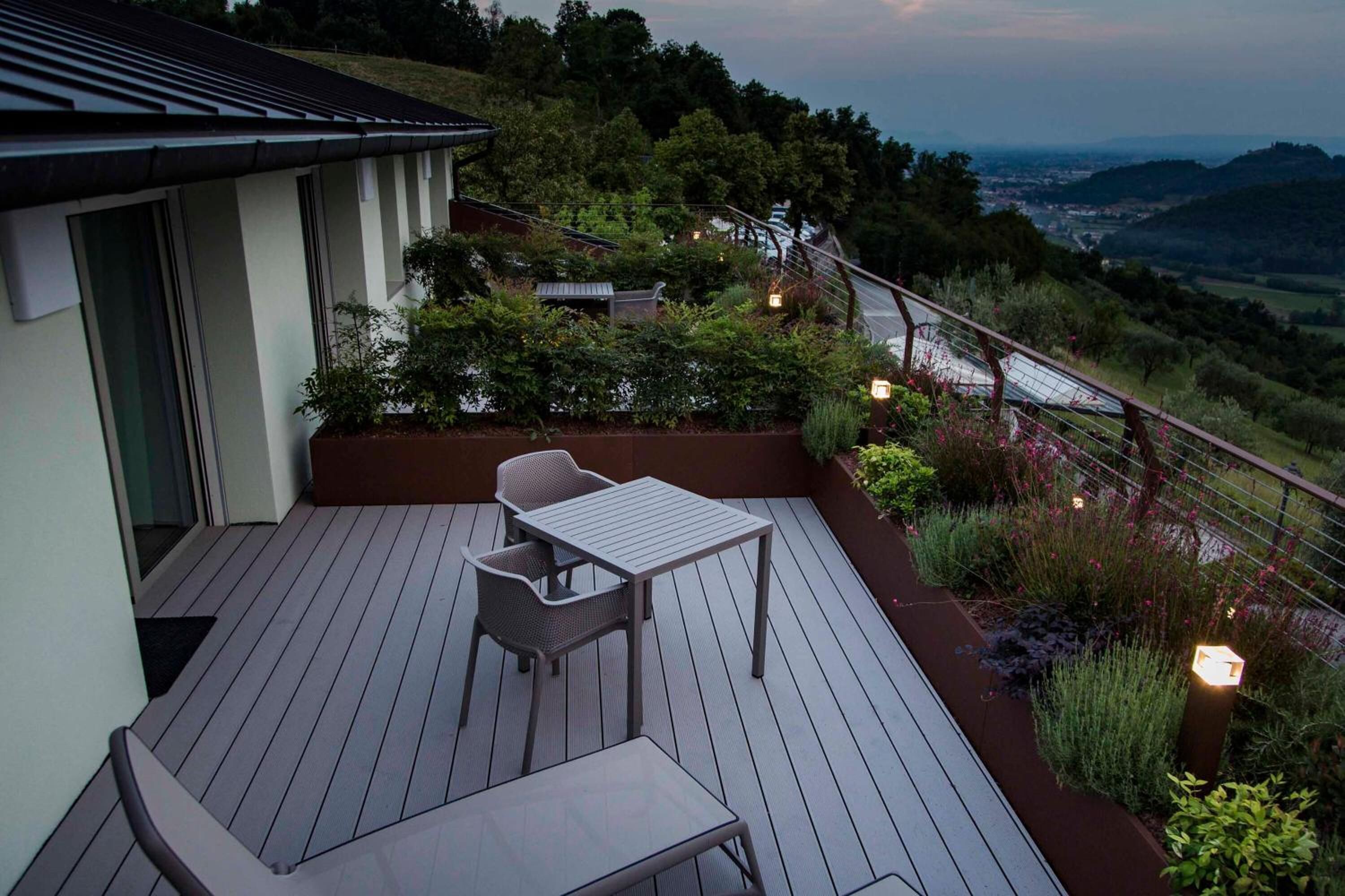 A wooden deck with a table and chairs, surrounded by greenery and overlooking a scenic landscape.