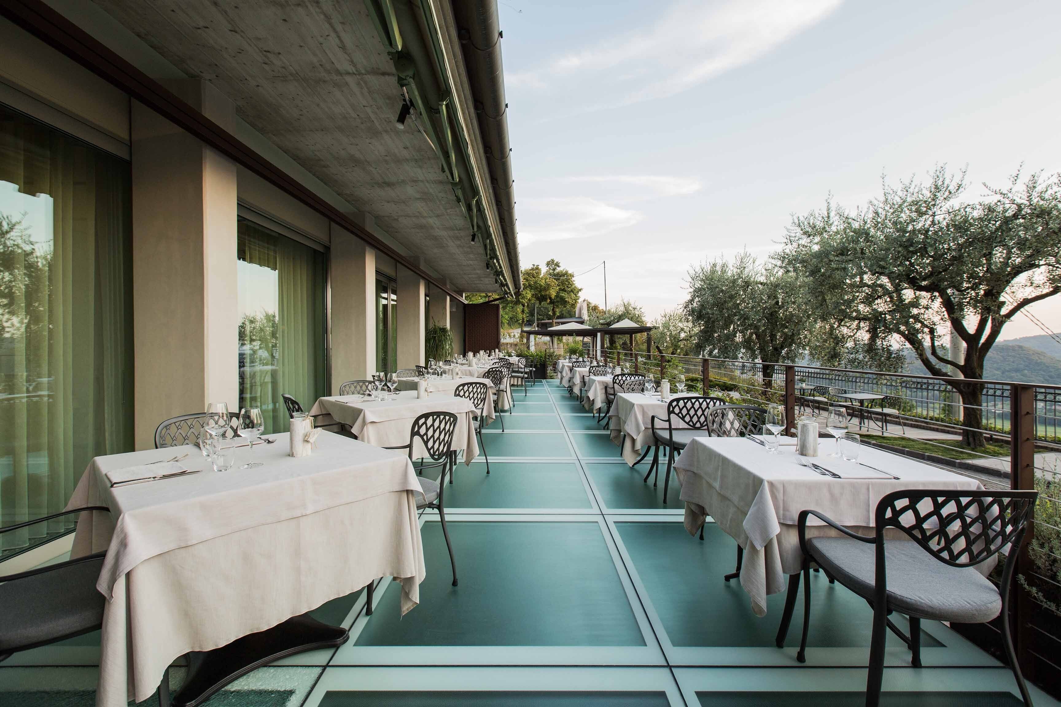 Un restaurante con terraza, mesas dispuestas con manteles blancos y sillas, y vistas a árboles y una montaña al fondo.