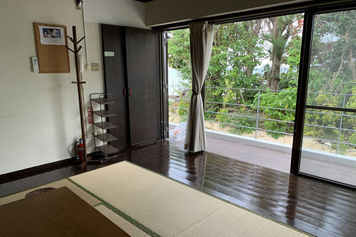 A room with a wooden table, a fire extinguisher, a shelf, and a window with a view of greenery.