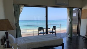 Een hotelkamer met een bed, een bureau met een lamp en uitzicht op het strand en de oceaan door grote ramen.