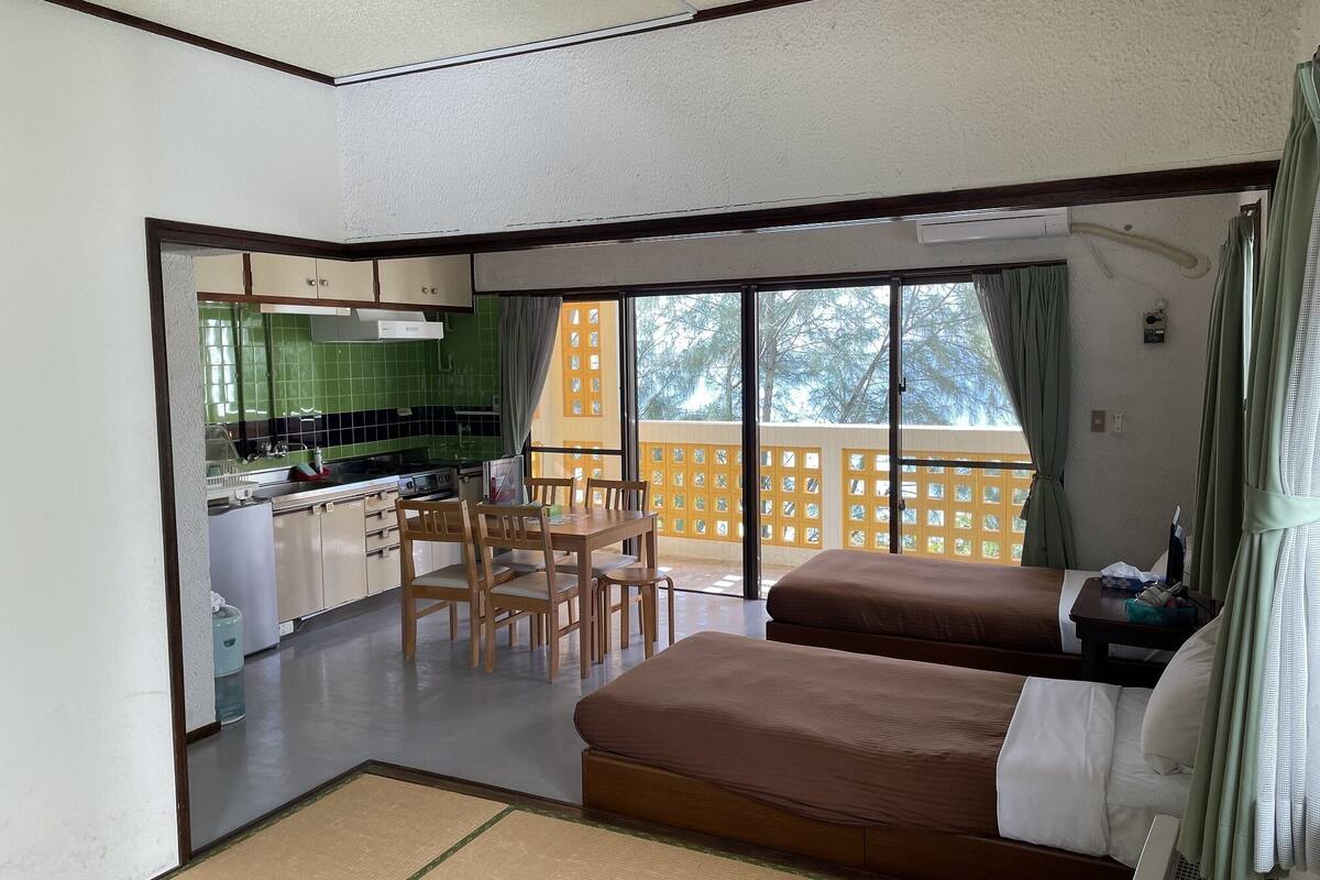 A room with a kitchenette, a dining table with chairs, two beds, and a view of a wooden deck through large windows.