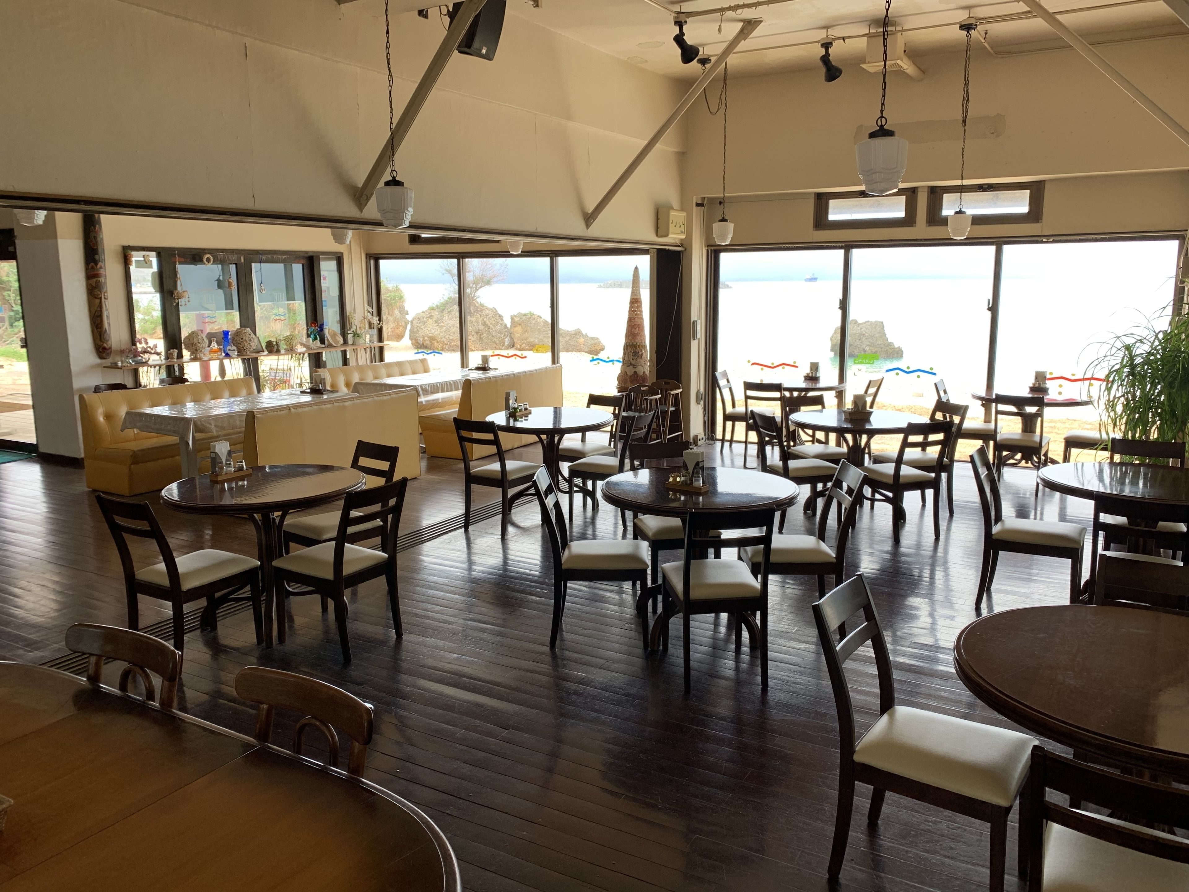 Un comedor amplio con varias mesas y sillas, ventanas grandes que ofrecen vistas al mar, y una zona de bar con mostrador y taburetes.