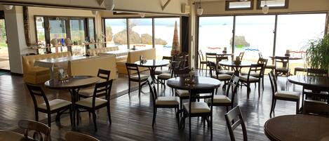 Un comedor amplio con varias mesas y sillas, ventanas grandes que ofrecen vistas al mar, y una zona de bar con mostrador y taburetes.