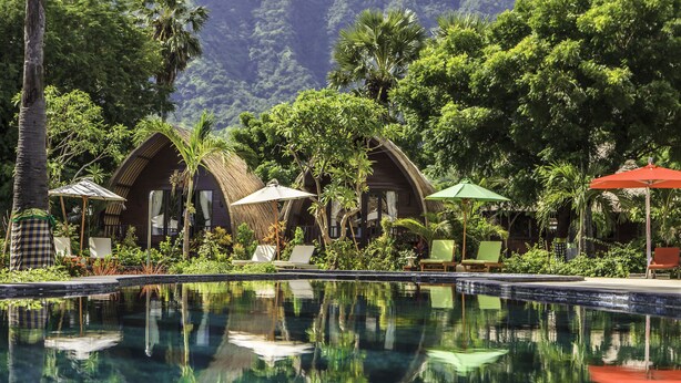 Una piscina con sillas de descanso y sombrillas, rodeada de exuberante vegetación y cabañas con techo de paja.