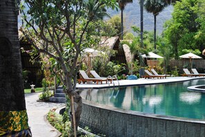 Zona de piscina con sillones de descanso y sombrillas, rodeada de vegetación tropical y un muro de piedra.