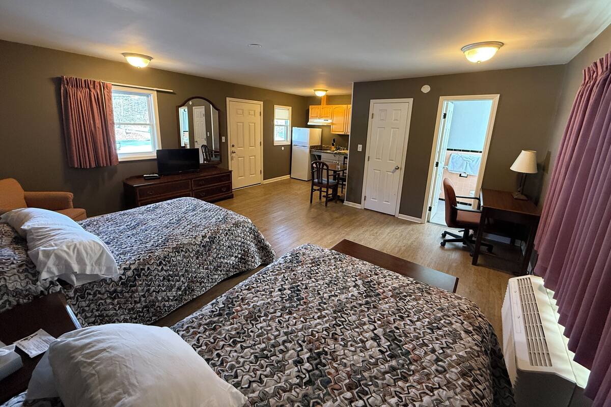 A hotel room with two beds, a desk, a chair, a TV, and a kitchenette.