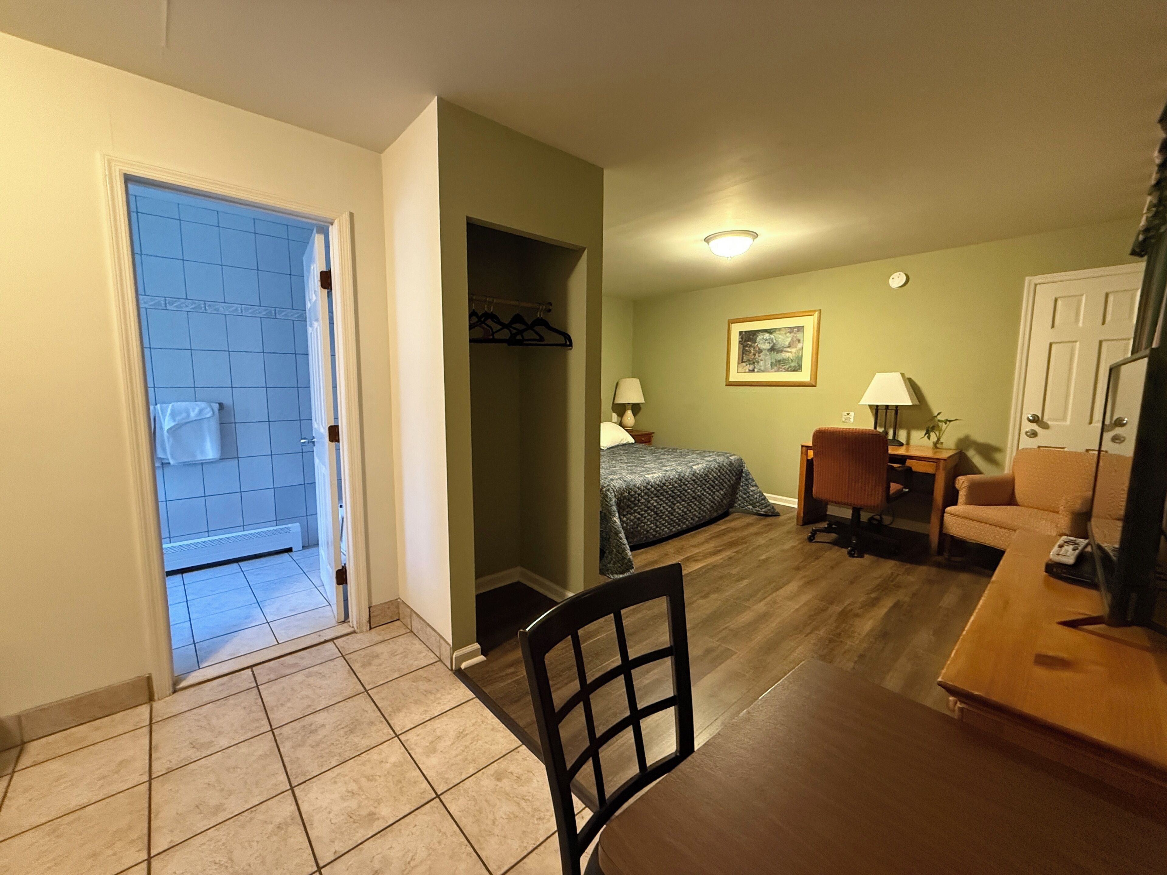 Quarto de hotel com cama, secretária, cadeira, abajur, cabideiro e porta para um banheiro.