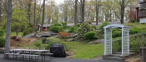 A picnic area with benches and a covered grill, surrounded by trees and a small garden with a white gazebo.