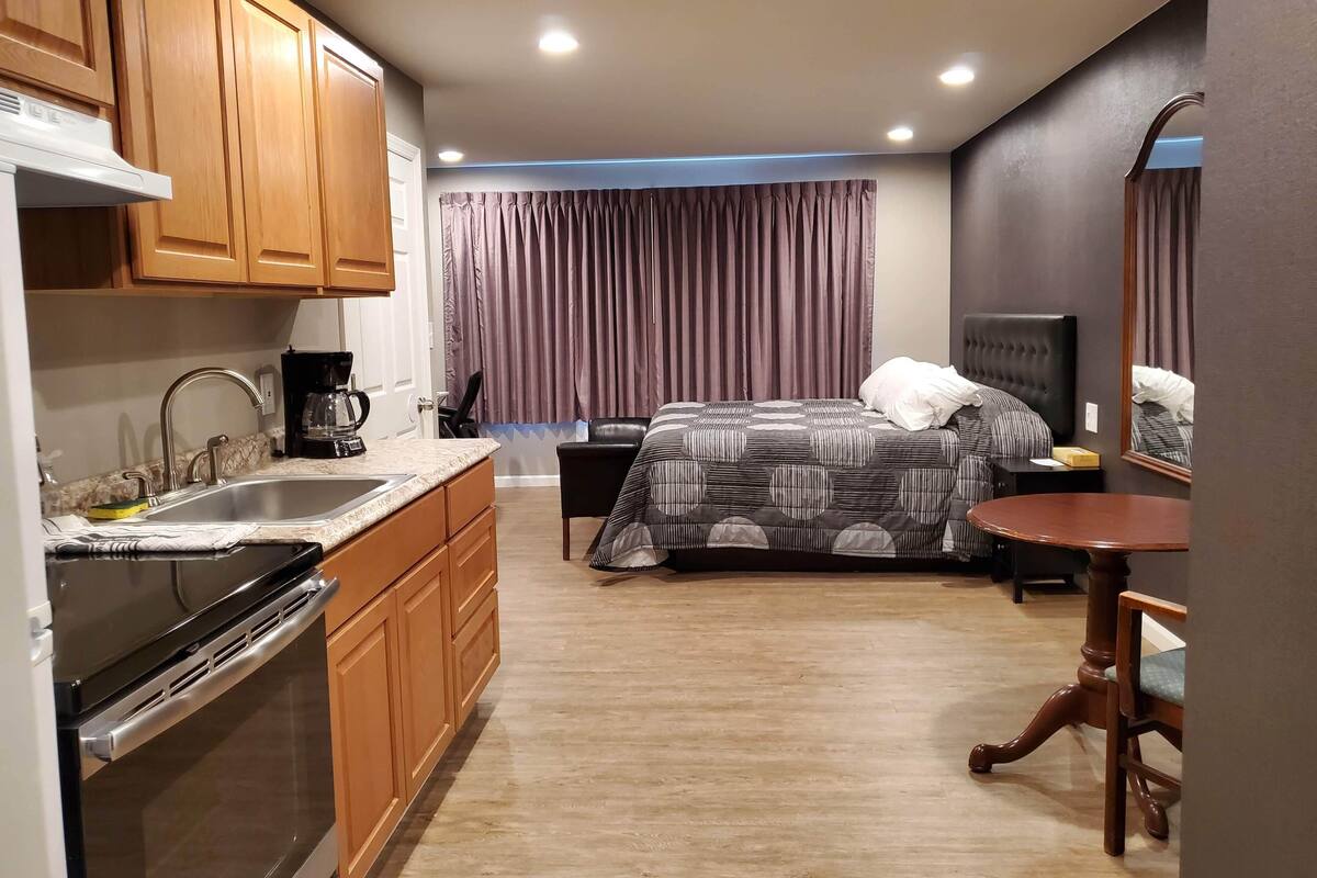 A compact hotel room with a kitchenette, a bed with a patterned bedspread, a bedside table, and a chair.