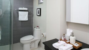 A modern bathroom with a glass shower enclosure, a toilet, a sink, and a vanity with toiletries. - The Grand Suree Residence (Bangkok)