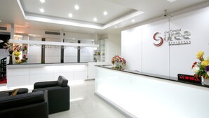 A modern reception area with a white reception desk, black sofas, and a digital clock displaying the time. - The Grand Suree Residence (Bangkok)