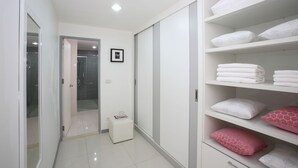A modern bathroom with a glass shower enclosure, a white vanity, and a neatly organized shelf with towels and pillows. - The Grand Suree Residence (Bangkok)
