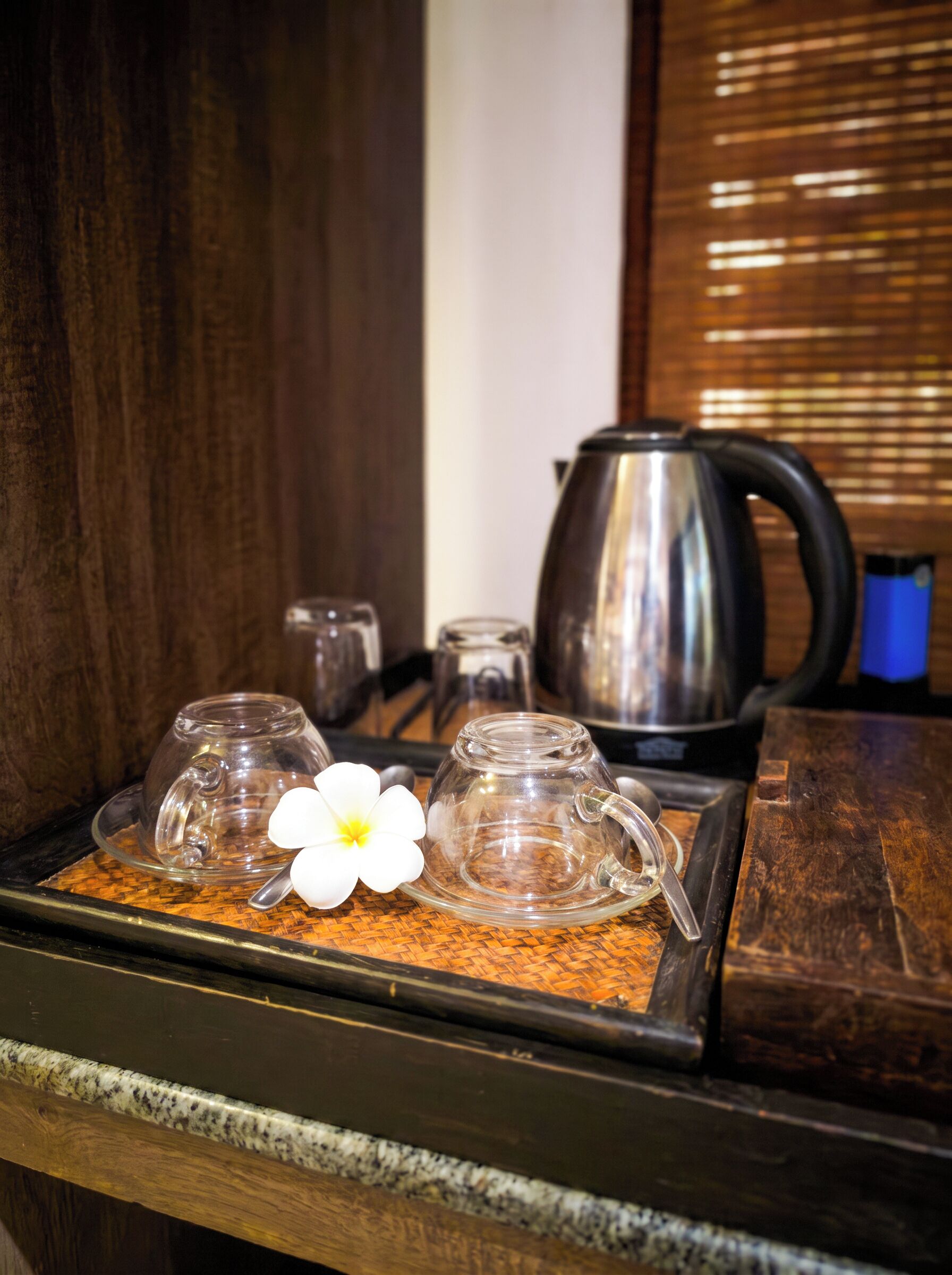 A tray with a glass teapot, two glass cups, and a white flower on a woven mat.