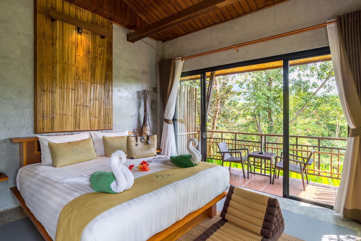 A spacious bedroom with a large bed, a balcony with a table and chairs, and a view of greenery.