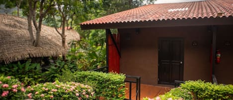 Un pequeño edificio con tejado de baldosas, una puerta y una ventana. Se observa una estructura de techo de paja y vegetación en el fondo.