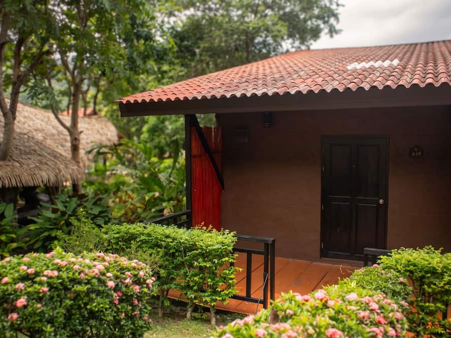 Un pequeño edificio con tejado de baldosas, una puerta y una ventana. Se observa una estructura de techo de paja y vegetación en el fondo.