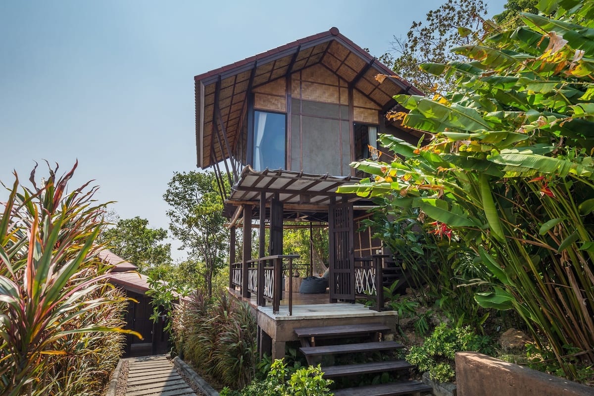 A two-story wooden structure with a balcony, surrounded by lush greenery and plants.