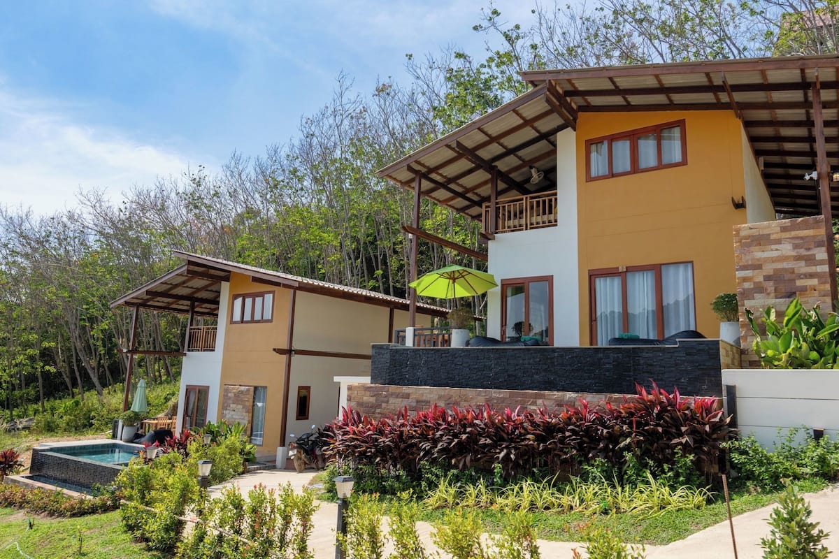 A two-story house with a balcony, a swimming pool, and a garden with various plants.