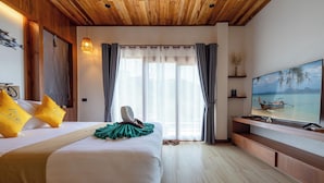 A hotel room with a wooden ceiling, a large bed with yellow pillows, a green throw, a TV on a wooden stand, and a view of the sea through the window. - Alama Sea Village Resort (Ko Lanta)