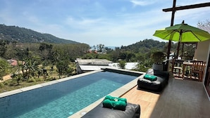 A poolside area with a lounge chair and a green towel, a patio with a table and chairs, and a view of a hilly landscape. - Alama Sea Village Resort (Ko Lanta)