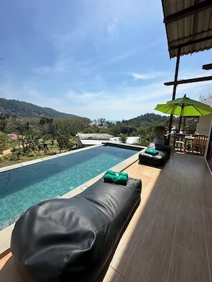 A poolside area with a lounge chair and a green towel, a patio with a table and chairs, and a view of a hilly landscape. - Alama Sea Village Resort (Ko Lanta)