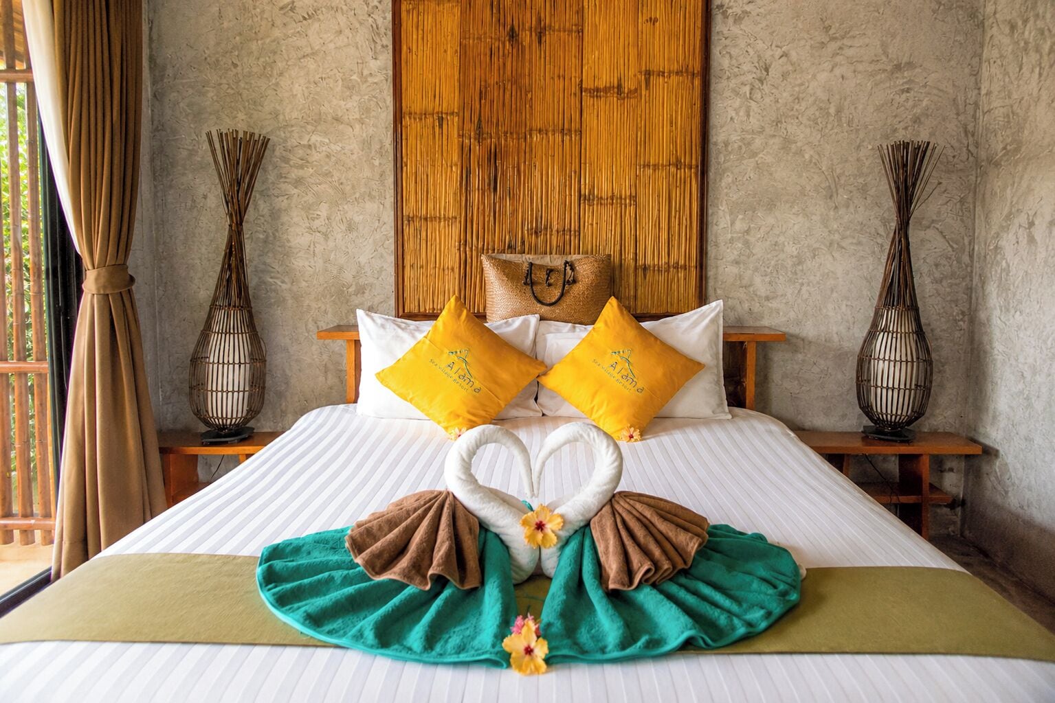 Una cama con una toalla dispuesta en forma de corazón, dos almohadas amarillas y una manta verde.
