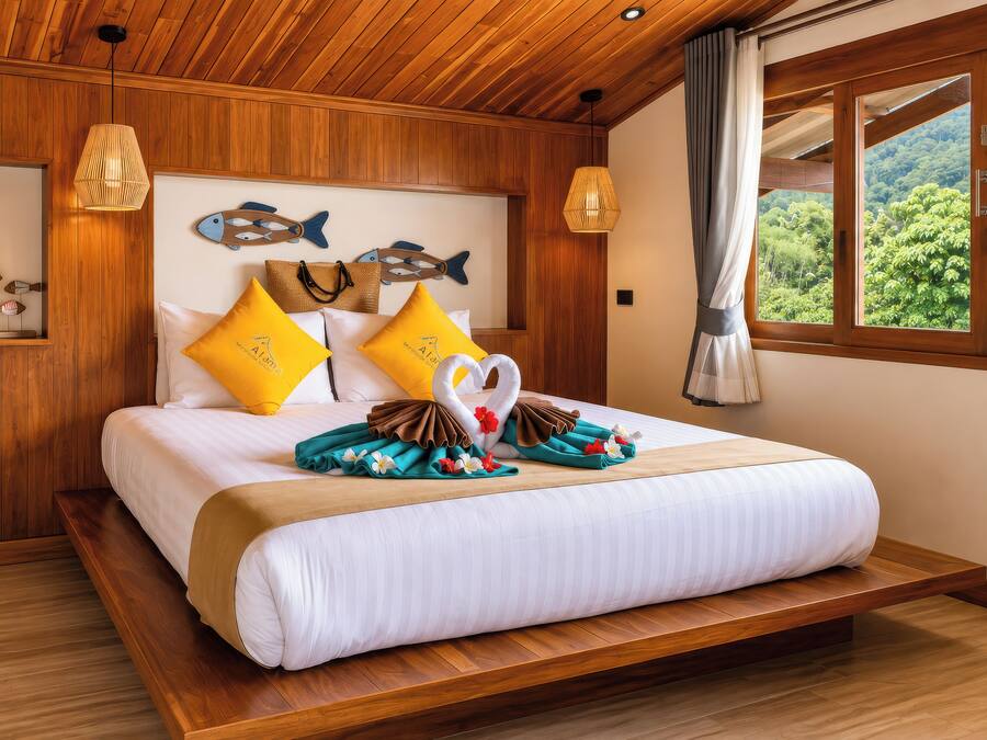 Habitación de hotel con techo de madera, cama con ropa blanca, almohadas amarillas y vista a un bosque por la ventana.