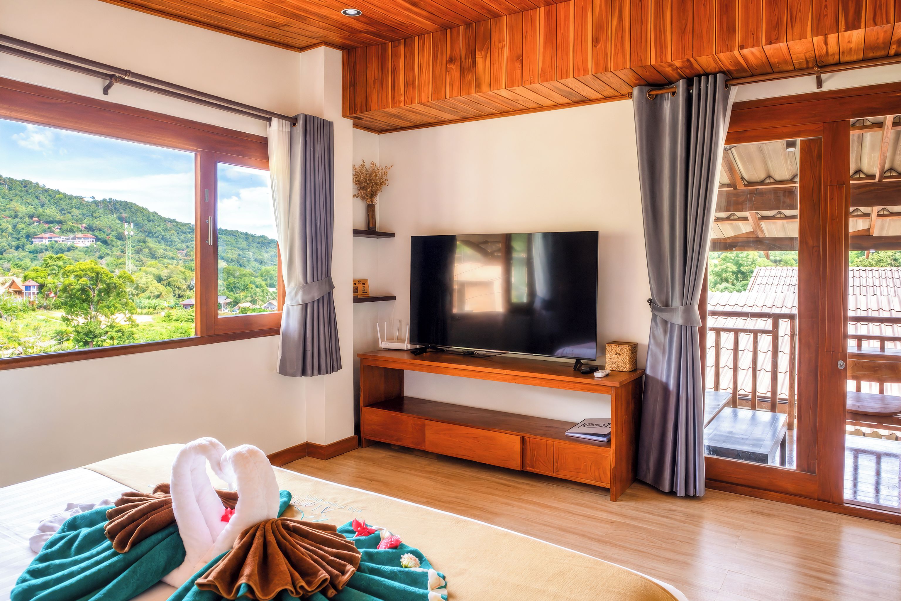 A hotel room with a large TV, wooden furniture, a bed with a decorative swan, and a balcony with a view of the hills.