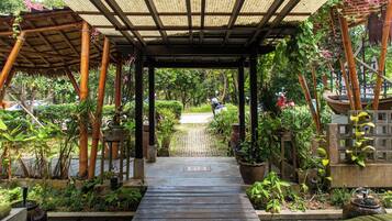 A covered walkway with a wooden floor leading to a path, surrounded by lush greenery and potted plants.
