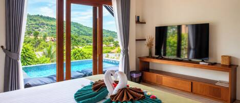A hotel room with a large bed, a television on a wooden stand, and a view of a swimming pool and greenery.