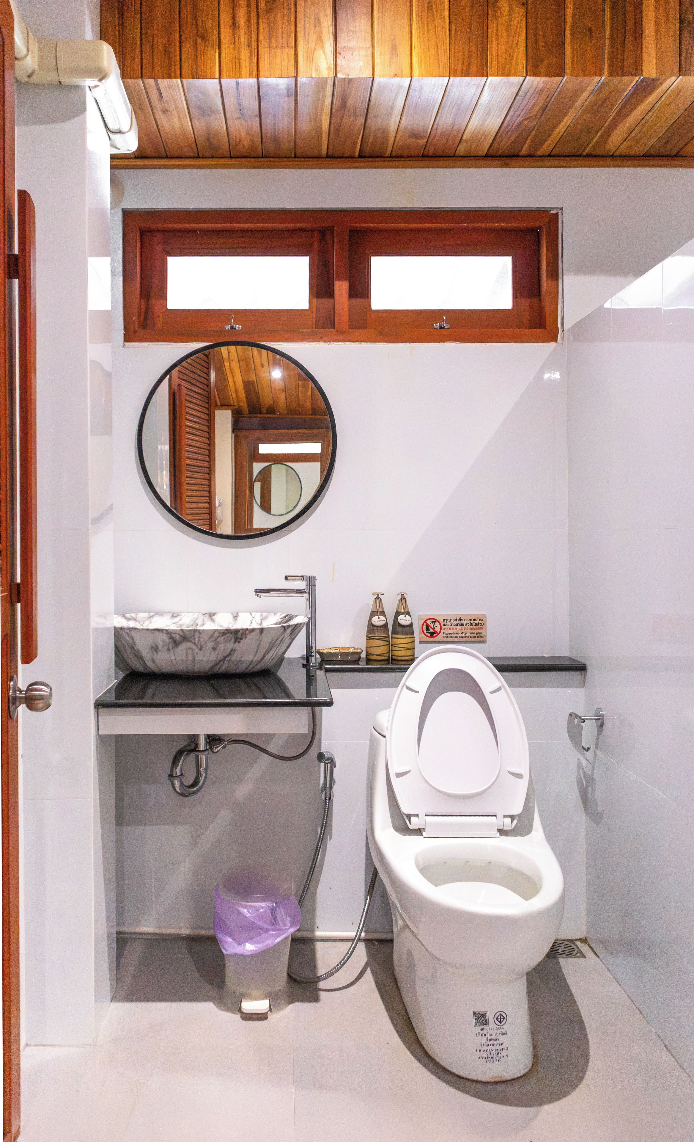 Une salle de bain avec des toilettes blanches, un lavabo doté d’un receveur en marbre, un meuble-lavabo en bois et un miroir au-dessus du lavabo.