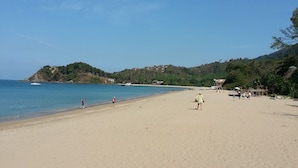 Una amplia playa de arena con agua azul cristalina, algunas personas caminando y colinas al fondo.