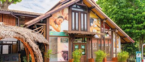 Un bâtiment traditionnel avec un toit de chaume, une grande affiche faisant la publicité de services de spa et un panneau indiquant les différentes prestations proposées, comme des massages et des soins d’aromathérapie.